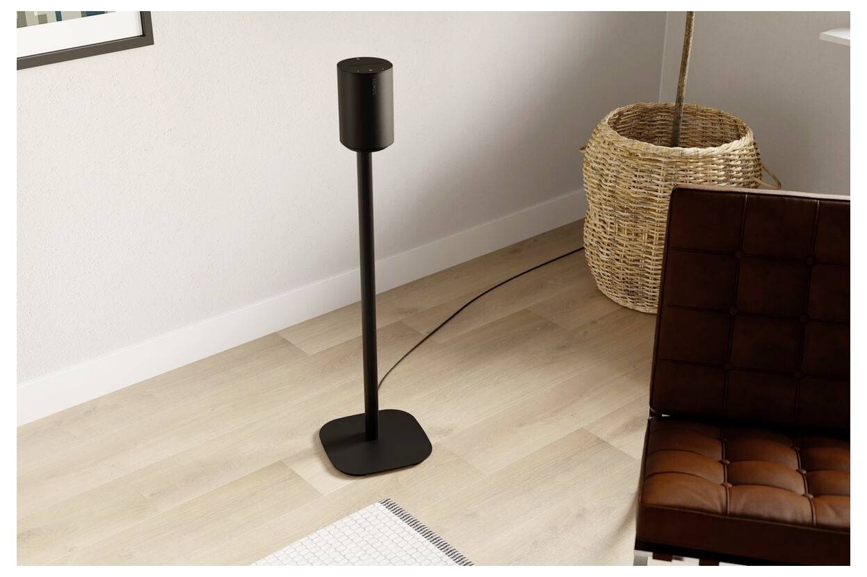 Moderní černý sloupový reproduktor v minimalistickém pokoji s proutěným košem a hnědou koženou židlí na světlé dřevěné podlaze.