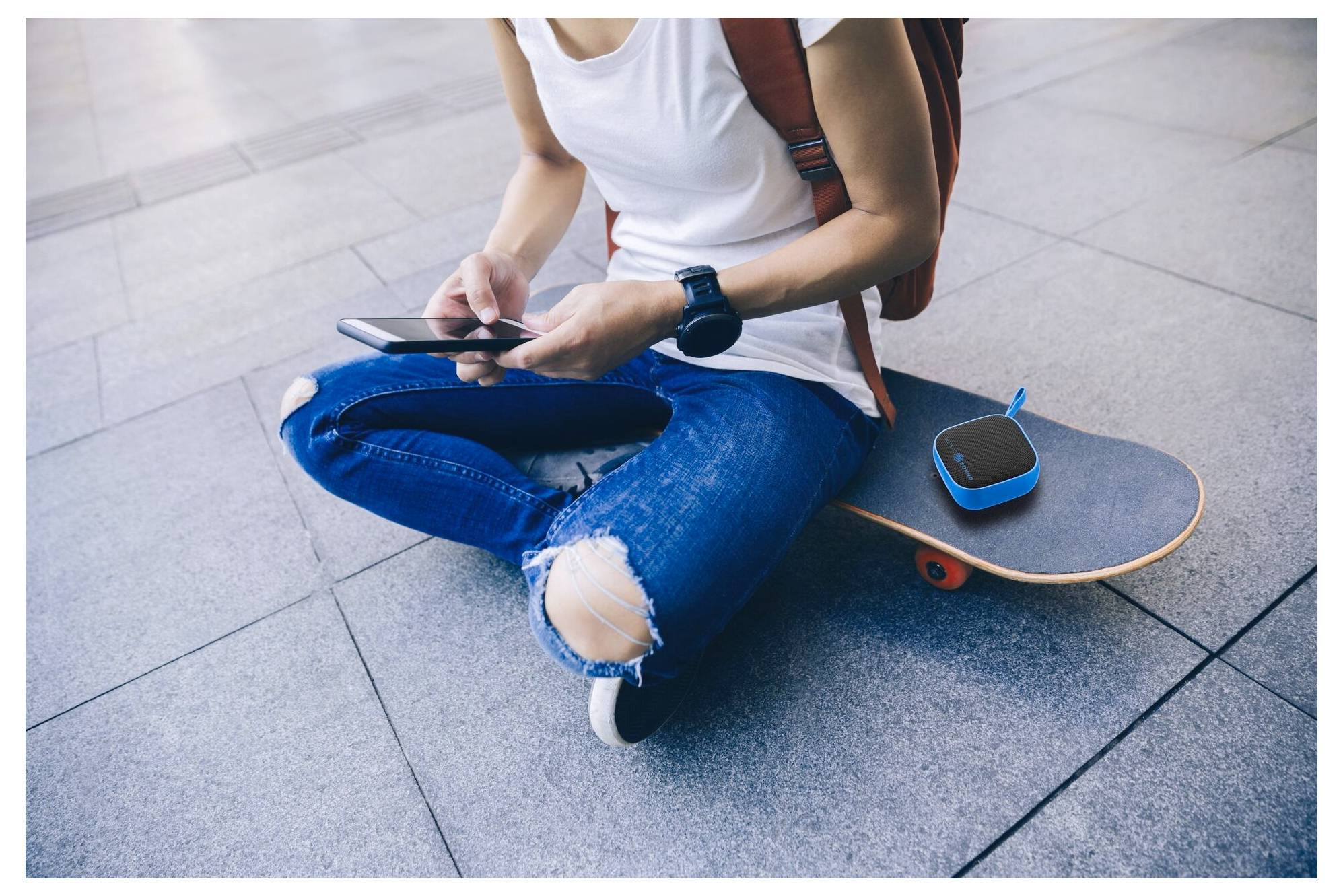 Osoba sedící na zemi se zkříženýma nohama na chodníku, používající chytrý telefon, s skateboardem a přenosným reproduktorem v blízkosti, což naznačuje relaxaci.