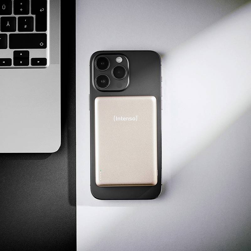 Smartphone s trojitým fotoaparátem leží na stole vedle klávesnice notebooku, nahoře je zlatá přenosná powerbanka označená „Intenso".
