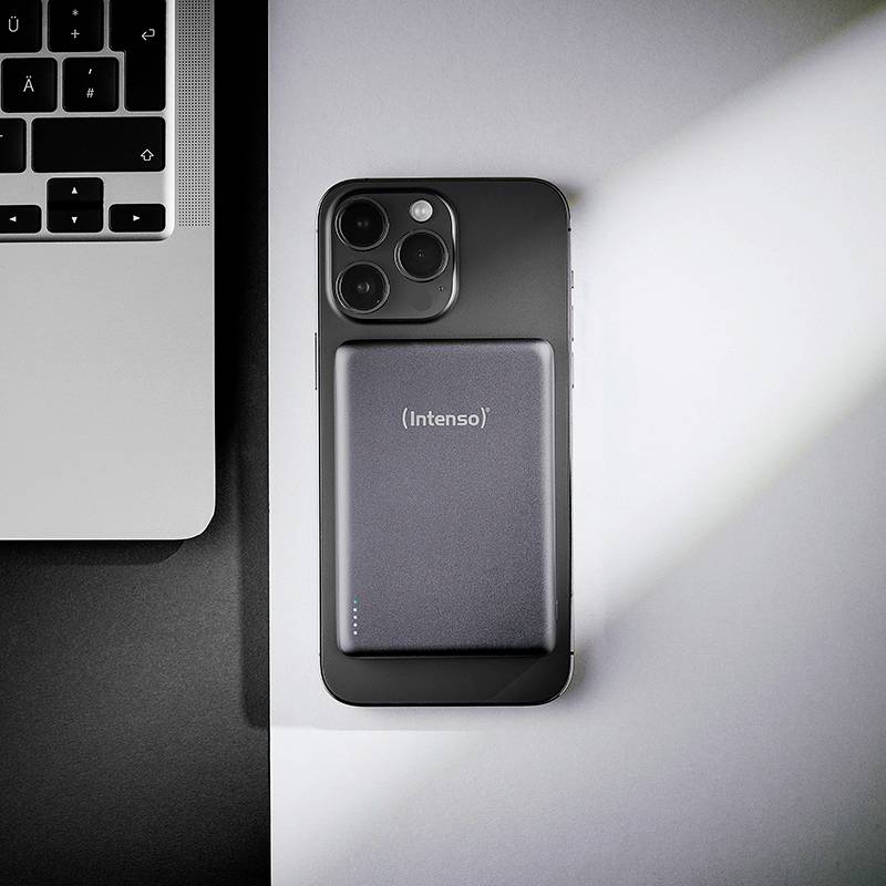 Smartphone na stole s obdélníkovou přenosnou powerbanka připojenou k jeho zadní straně. Část klávesnice notebooku je viditelná vlevo.
