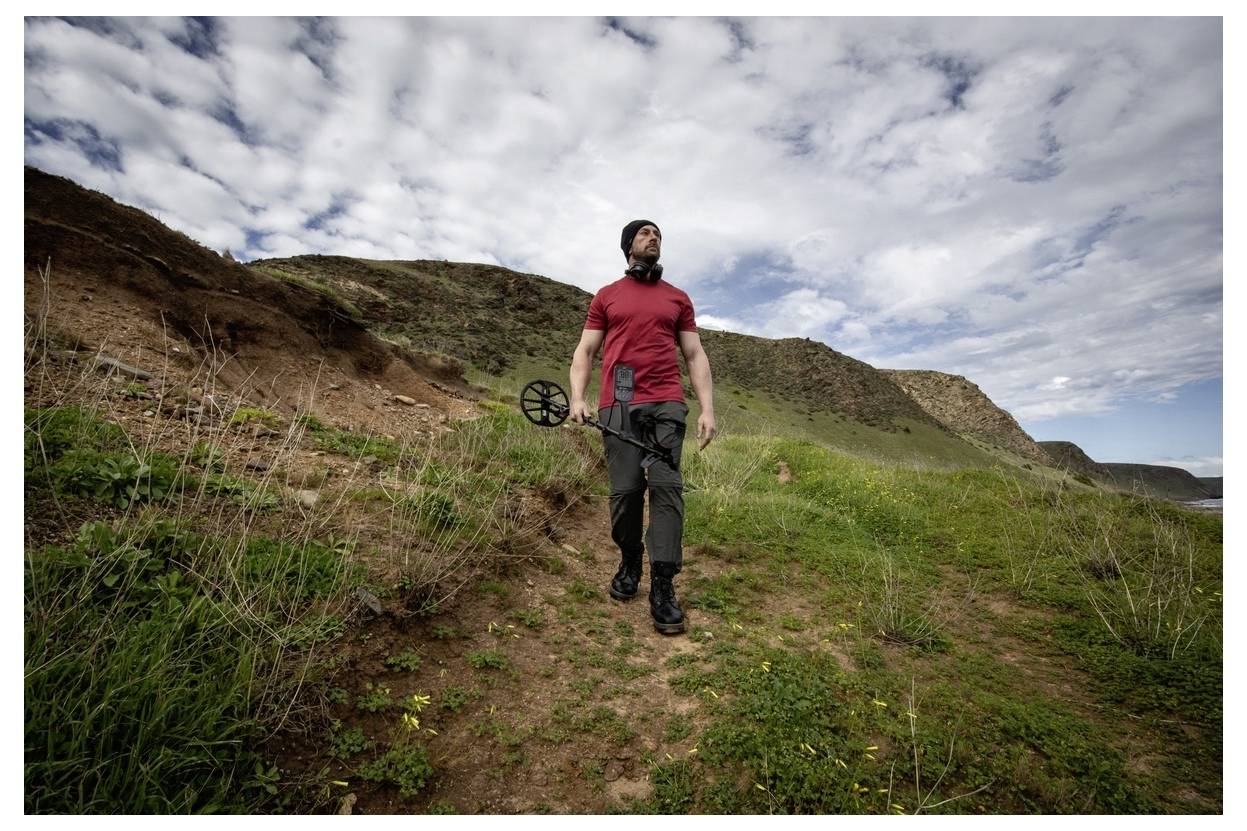 Osoba kráčející po travnatém kopci pod částečně zataženou oblohou, držící kovový detektor, oblečená v červené košili a černých kalhotách.
