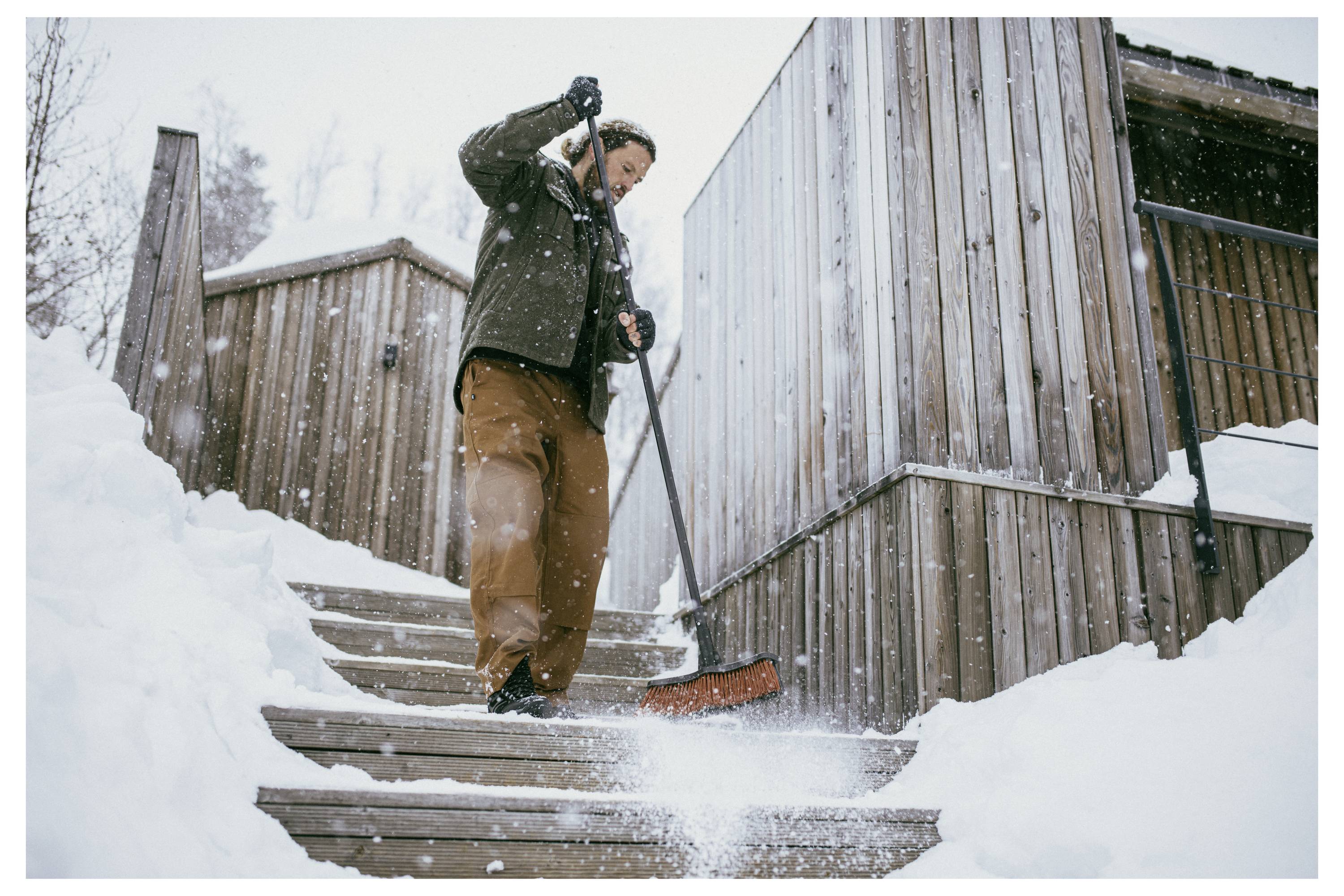 Osoba odhazuje sníh na dřevěných schodech venku u budovy v zasněžený den, oblečená v zimním kabátě a hnědých kalhotách.