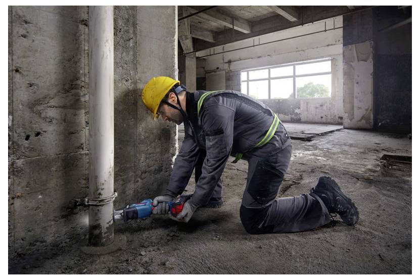 Dělník ve stavebním ochranném oděvu klečí na betonové podlaze a používá elektrický nástroj k práci na svislém potrubí ve velké, nedokončené místnosti.