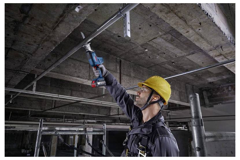 Dělník v žluté ochranné přilbě používá elektrický nástroj k instalaci kovových trubek na betonový strop, což naznačuje probíhající stavební práce.