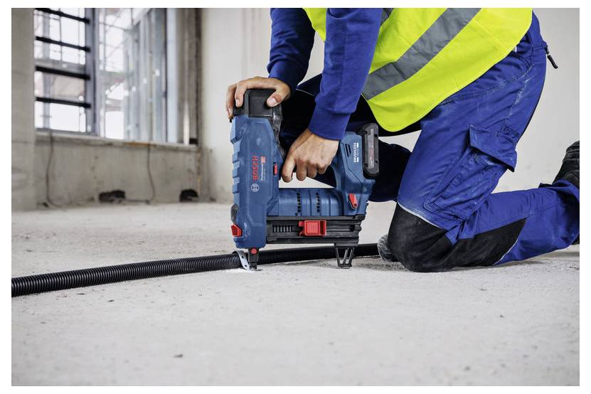 Stavební dělník v modré montérkové kombinéze a reflexní bezpečnostní vestě klečí na betonové podlaze a používá průmyslový nástroj pro upevňování nebo zajišťování materiálů.