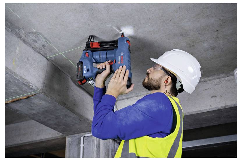Osoba v bezpečnostní vestě a helmě vrtá do betonového stropu pomocí elektrického nářadí, pracuje na stavbě nebo rekonstrukci.