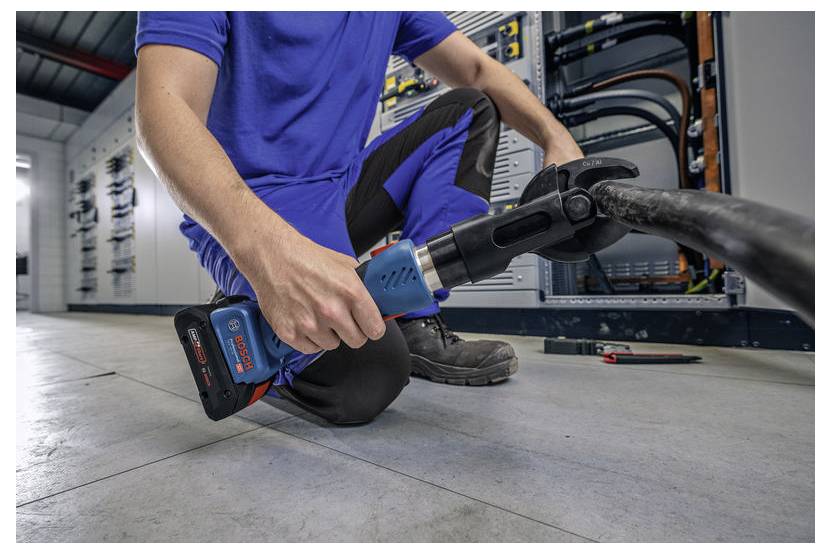 Dělník v modré uniformě používá modro-černý elektrický nástroj k rozříznutí tlustého kabelu v průmyslovém prostředí s elektrickými rozvaděči v pozadí.
