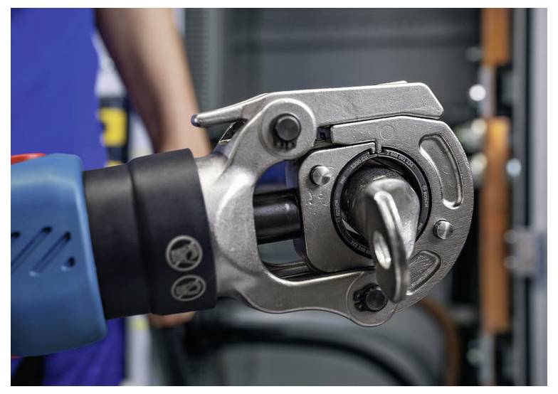 Detailní záběr hydraulického lisovacího nástroje, který lisuje kovovou objímku. V rozostřeném pozadí stojí osoba v modrém oblečení.