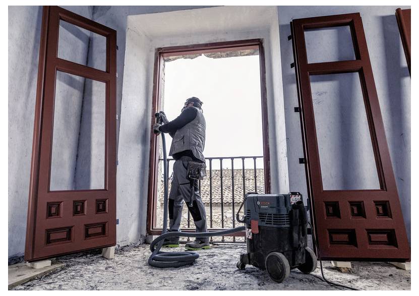 Osoba v ochranném oblečení používá elektrický nástroj k práci na balkónovém okně, přičemž demontované okenní rámy jsou opřené o stěny.