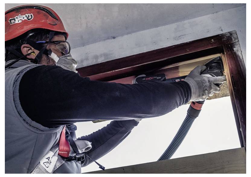 Dělník ve stavebním ochranném oděvu používá elektrický nástroj k řezání dřevěného rámu. Slunečního světlo osvětluje scénu zvenčí.