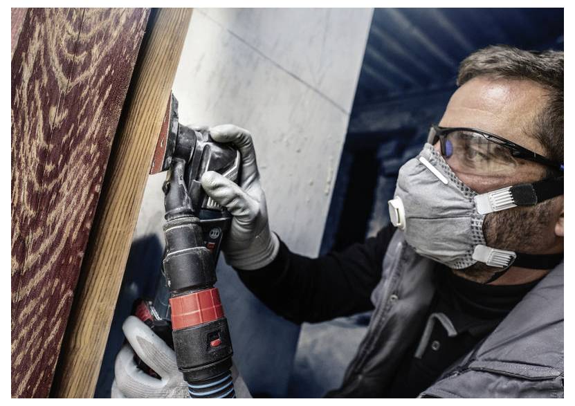 Osoba v ochranných pomůckách používá elektrické nářadí při práci na dřevěném povrchu.