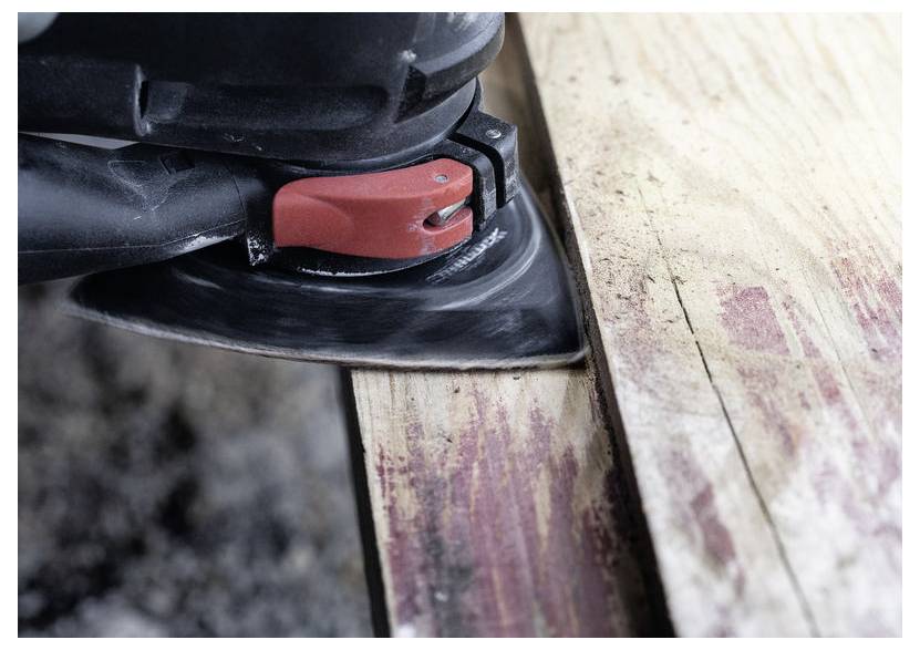 Detailní záběr elektrické brusky s červeným spínačem, která hladí hranu dřevěné desky. Dřevo vykazuje stopy po předchozím nátěru nebo opotřebení.