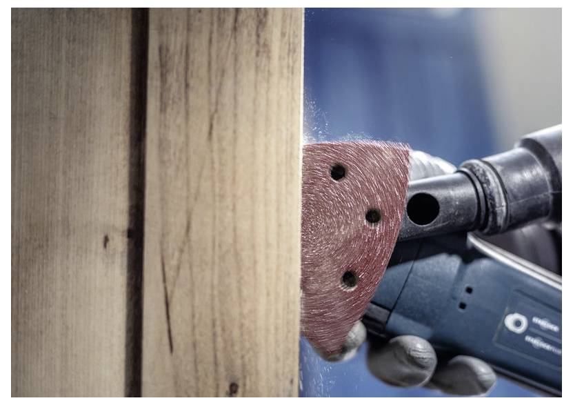 Detailní záběr ruky, která používá elektrickou brusku na dřevěném povrchu, vyhlazuje dřevní strukturu. Ve vzduchu jsou viditelné prachové částice.