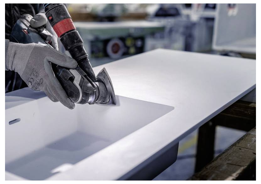 Osoba v rukavicích používá elektrický nástroj k leštění hran bílé pracovní desky se zabudovaným dřezem.