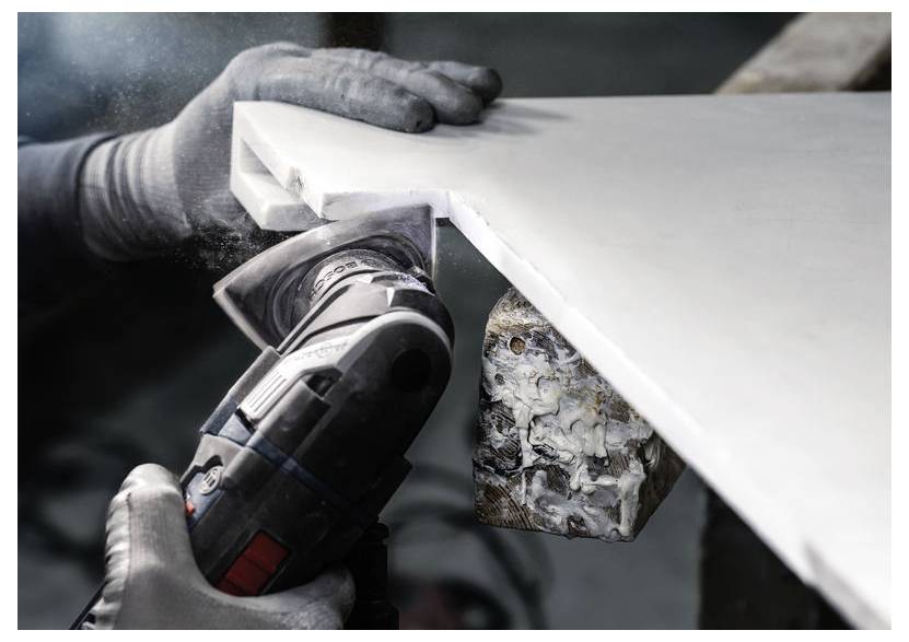 Osoba v rukavicích používá elektrický nástroj k broušení hrany velké bílé plochy, přičemž se zaměřuje na roh. Jsou viditelné prachové částice.