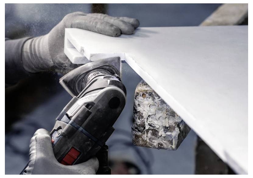 Osoba v rukavicích používá elektrický nástroj k broušení hrany bílé kamenné desky, přičemž vytváří prach. Svěrka drží desku pevně na místě.