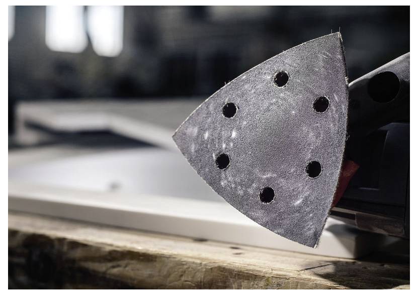 Detailní záběr hlavy brusky s připevněným brusným kotoučem, který má několik otvorů. Na pozadí je rozmazané pracovní prostředí.