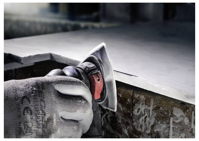 Rukavicí opatěná ruka drží elektrickou brusku, která vyhlazuje hranu dřevěného povrchu. Kolem pohybu nástroje jsou viditelné prachové částice.