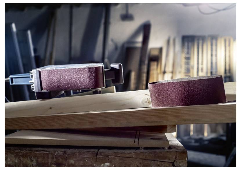 Pásová bruska a brusný pás na dřevěné desce v dílně s rozostřeným pozadím zobrazujícím různé dřevěné desky.