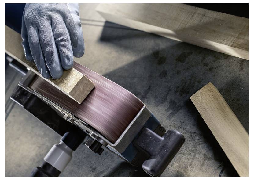 Rukavicí chráněná ruka drží kus dřeva, který je broušen na pásové brusce, v blízkosti jsou další dřevěné kusy, což zvýrazňuje probíhající truhlářskou práci.