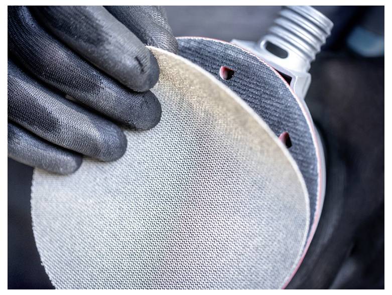 Osoba v černých rukavicích umisťuje kruhový smirkový papír na elektrickou brusku a připravuje se na broušení.