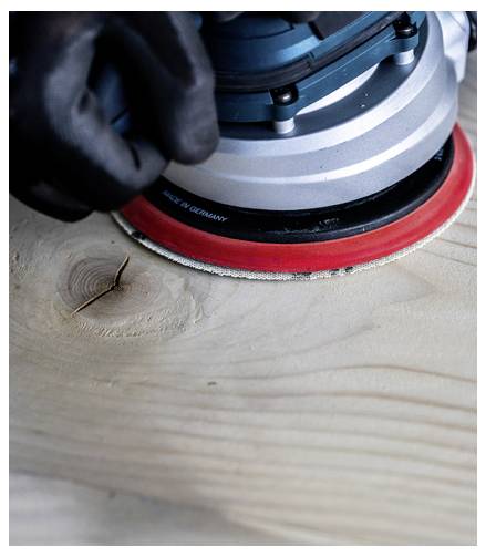 Osoba v rukavici používá kruhovou brusku na dřevěném povrchu a vyhlazuje strukturu dřeva.