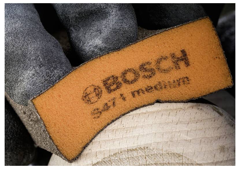 Rukavicí opatěná ruka drží oranžový brusný kotouč označený "BOSCH 347 medium" přiložený k dřevěnému povrchu, což naznačuje truhlářské práce nebo přípravu povrchu.