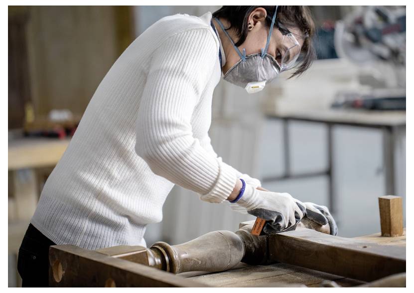 Osoba v ochranných brýlích a respirátoru brousí dřevěnou nohu v dílně a používá ochranné rukavice pro bezpečnost.