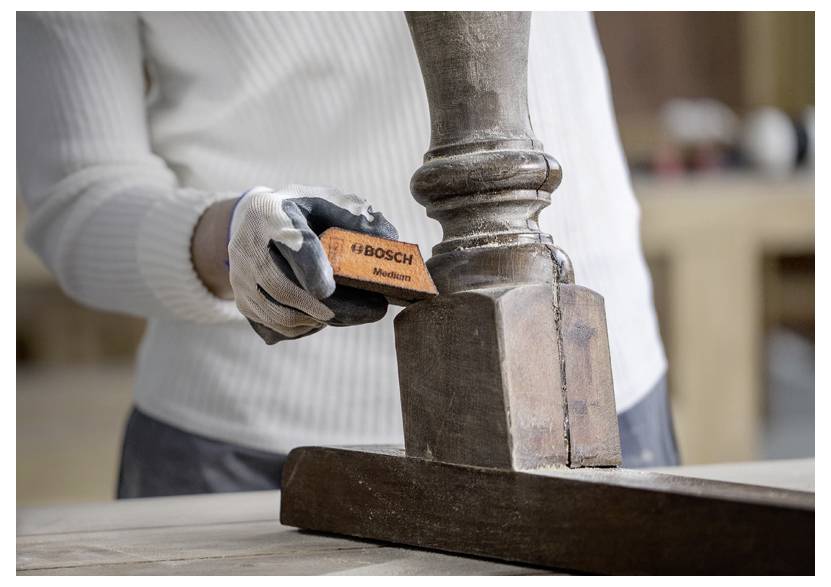 Osoba v rukavicích brousí dřevěnou nohu stolu pomocí brusného bloku značky Bosch v prostředí dílny.