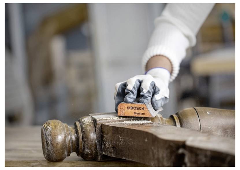 Osoba v rukavicích používá bruskou desku Bosch na staré noze židle a vyhlazuje povrch v prostředí dílny.