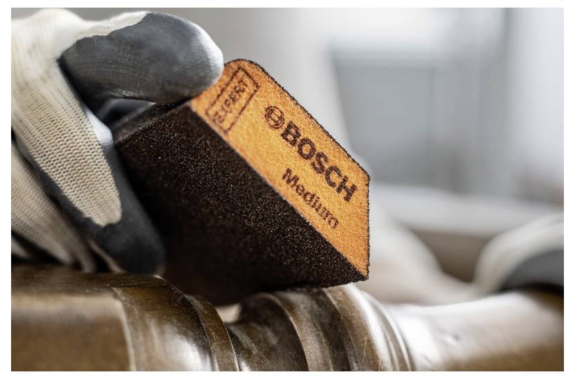 Rukavicí opatěná ruka drží střední brusnou houbu značky Bosch a vyhlazuje dřevěný povrch v truhlářské dílně.