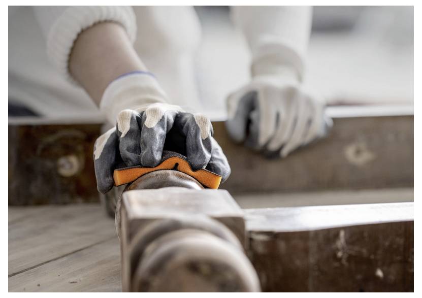 Osoba v ochranných rukavicích brousí dřevo oranžovým nástrojem a zaměřuje se na vyhlazení rohu, což naznačuje práci se dřevem nebo projekt pro kutily.