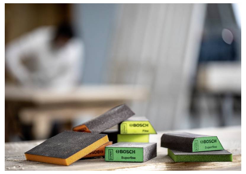 Na pracovním stole jsou umístěny brusné bloky označené 'Bosch Fine' a 'Bosch Superfine'. Osoba v pozadí je rozmazaná, což naznačuje probíhající práci.