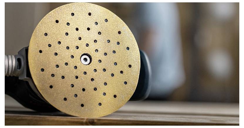 Detailní záběr kruhového brusného kotouče s otvory, připevněného k elektrickému nástroji, položeného na dřevěném povrchu. Pozadí je rozmazané.