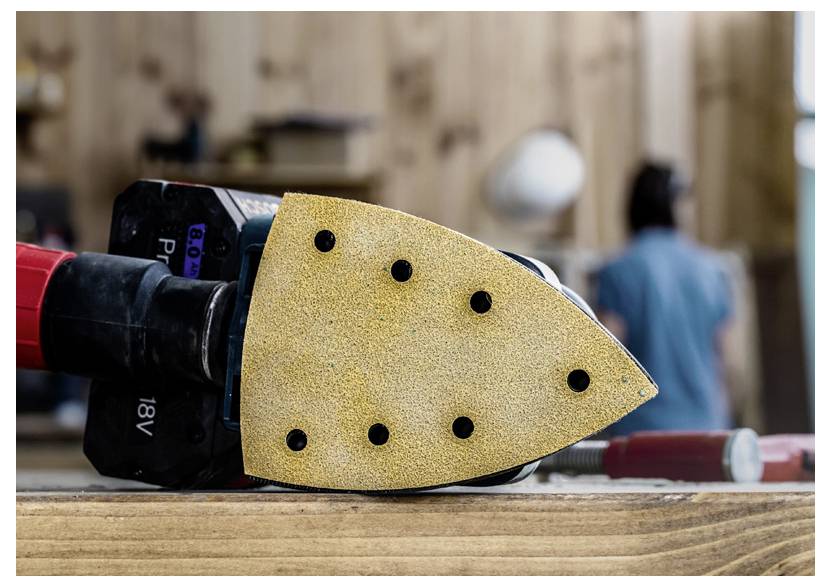 Detail záběru brusného nástroje s trojúhelníkovým brusným papírem připevněným na dřevěném povrchu. Rozostřené pozadí zobrazuje prostředí dílny.