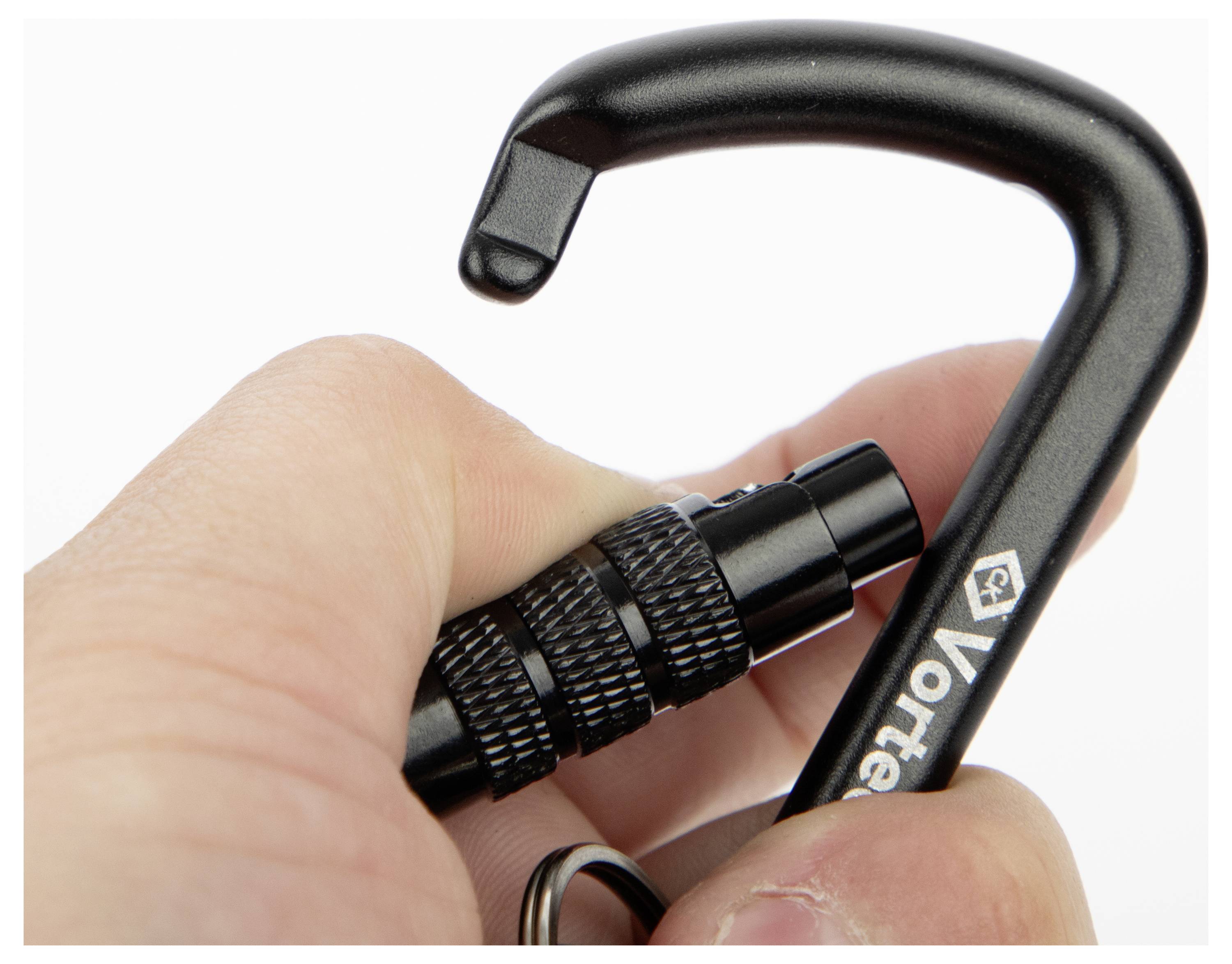 Ruka držící černou karabinu Vortex s uzamykacím mechanismem, částečně zakrývající některé prvky brandingu. Karabina má zakřivenou, otevřenou pojistku.