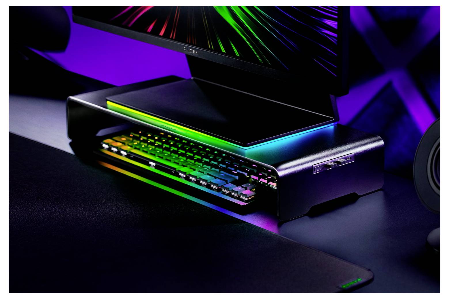 Monitor se zářícími neonovými světly na stojanu, klávesnice pod ním, zářící vícebarevnými světly v tmavém prostředí místnosti.