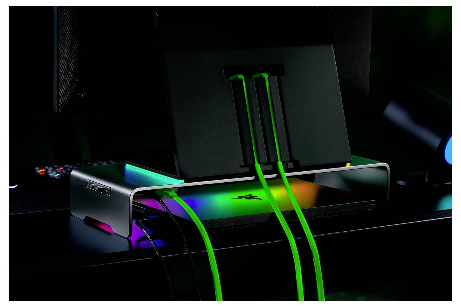 Elegantní laptop zavřený na stojanu s výraznými zelenými a modrými LED světly vyzařujícími z jeho povrchu, propojený zářícími zelenými kabely na tmavém pracovním stole.