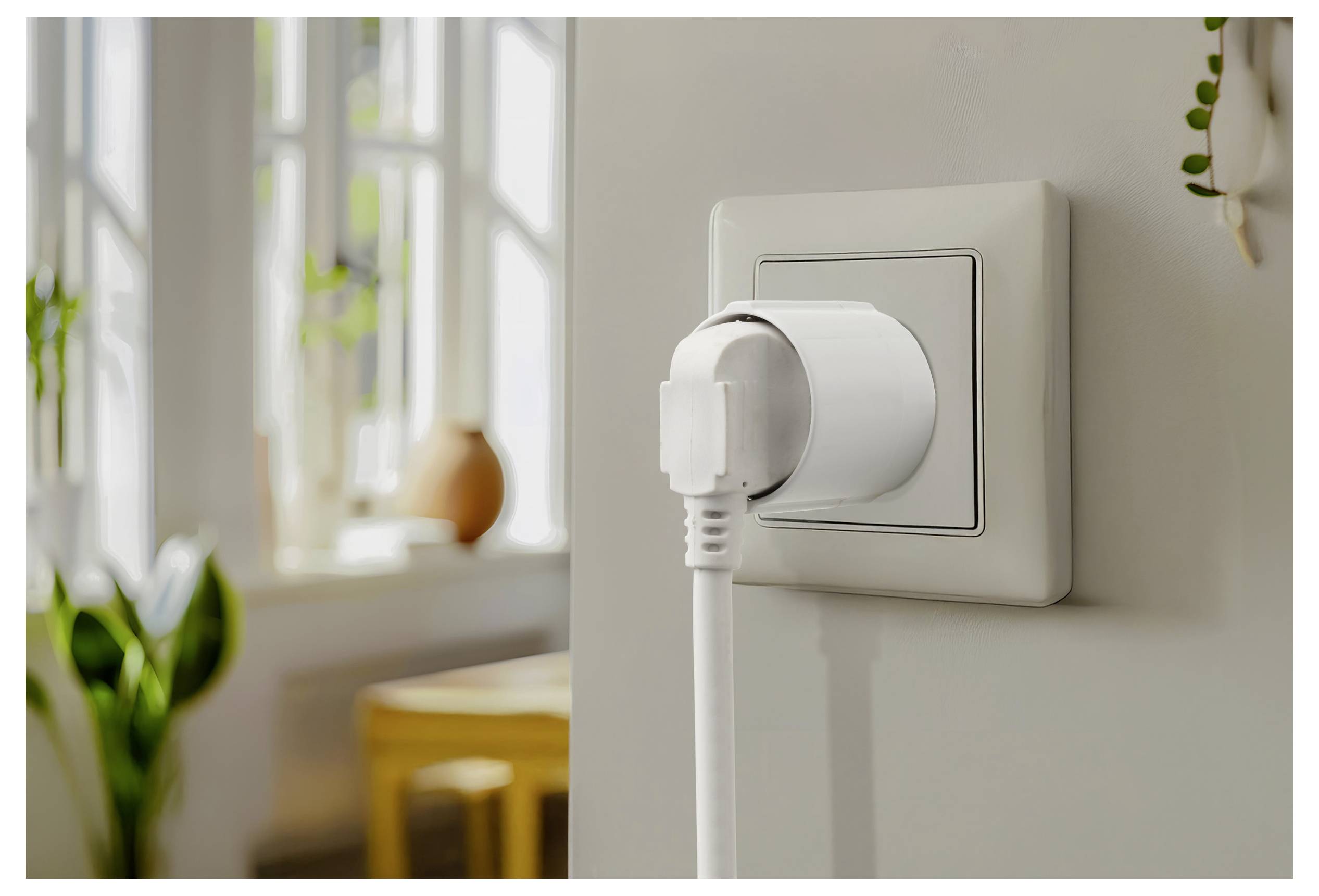Bílá napájecí zástrčka je zasunuta do elektrické zásuvky v jasně osvětlené místnosti s rozmazaným pozadím znázorňujícím stůl a okno.