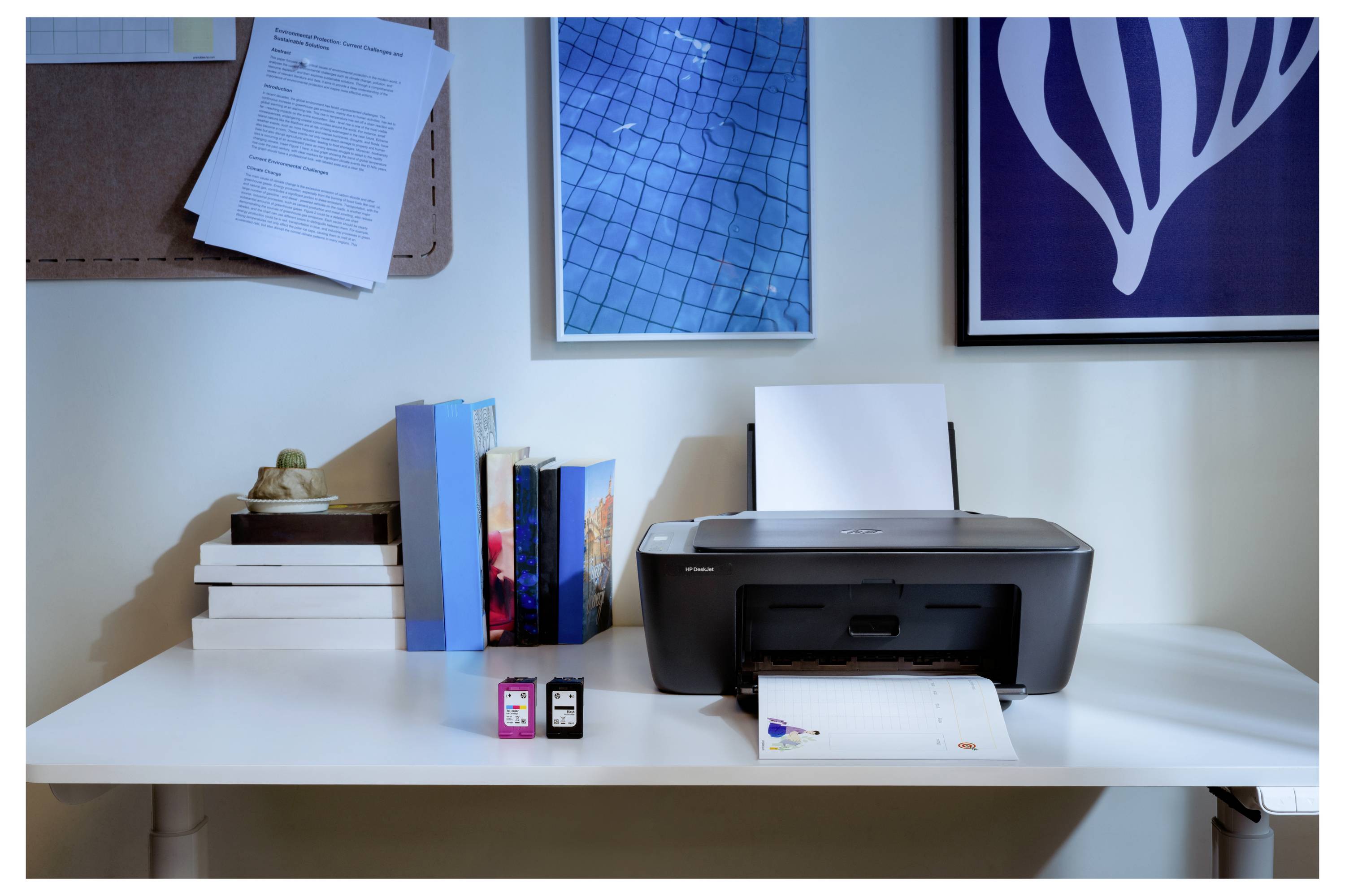 Nastavení domácího kancelářského stolu s tiskárnou, cartridgemi s inkoustem, naskládanými knihami a dekorativním nástěnným uměním v pozadí.