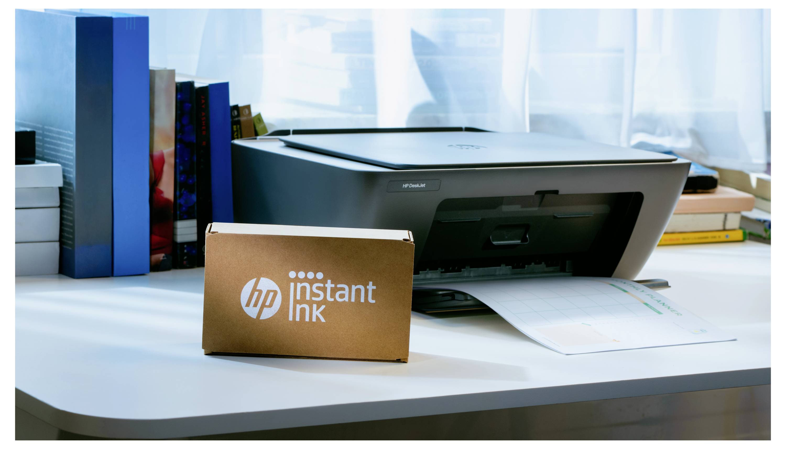 Tiskárna na stole vedle krabice označené "HP Instant Ink", z níž vychází vytištěná stránka. V pozadí jsou knihy a kancelářské pomůcky.