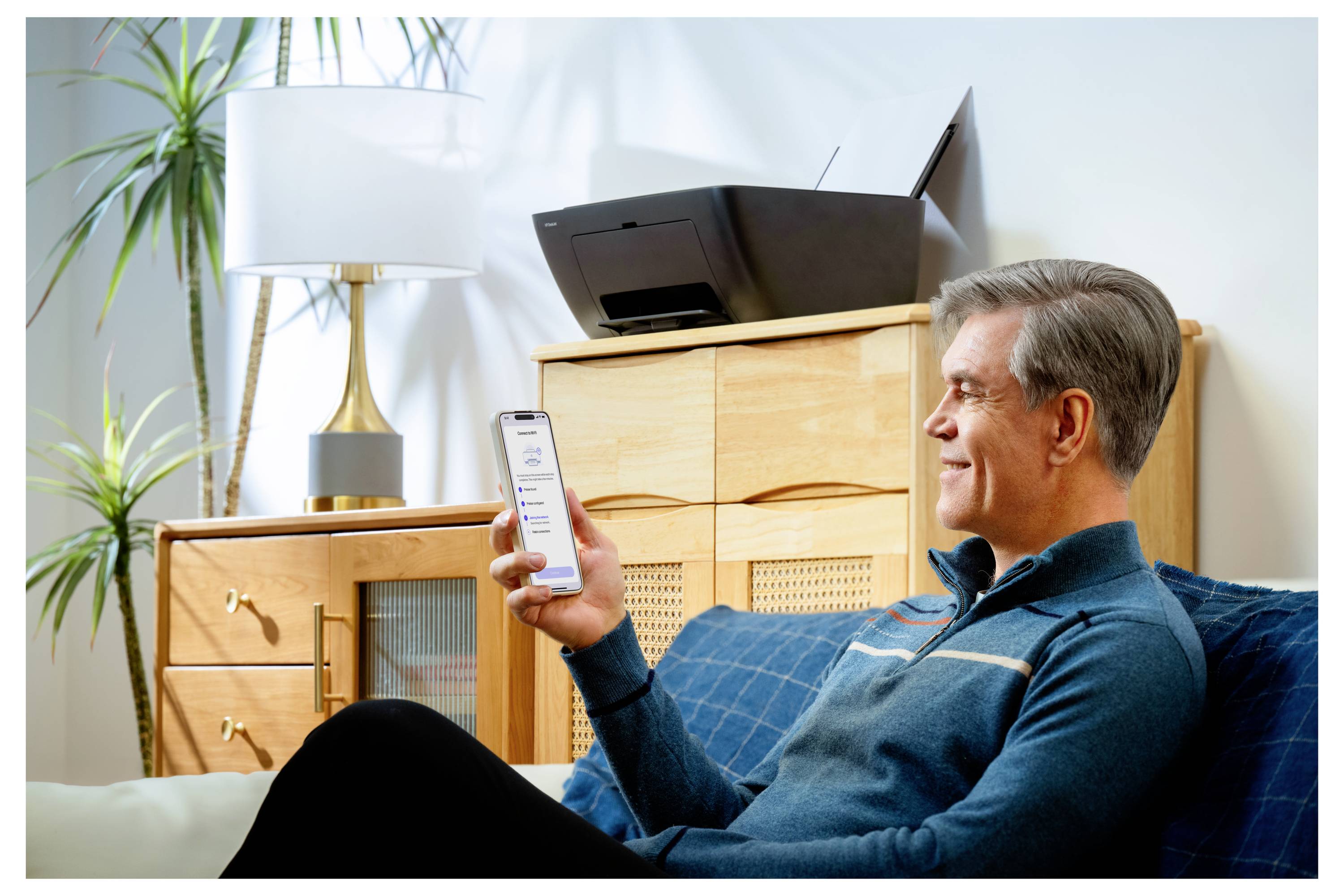 Muž sedící na pohovce, usmívající se při pohledu na chytrý telefon. V pozadí je viditelná multifunkční tiskárna. Prostředí je pohodlný obývací pokoj.