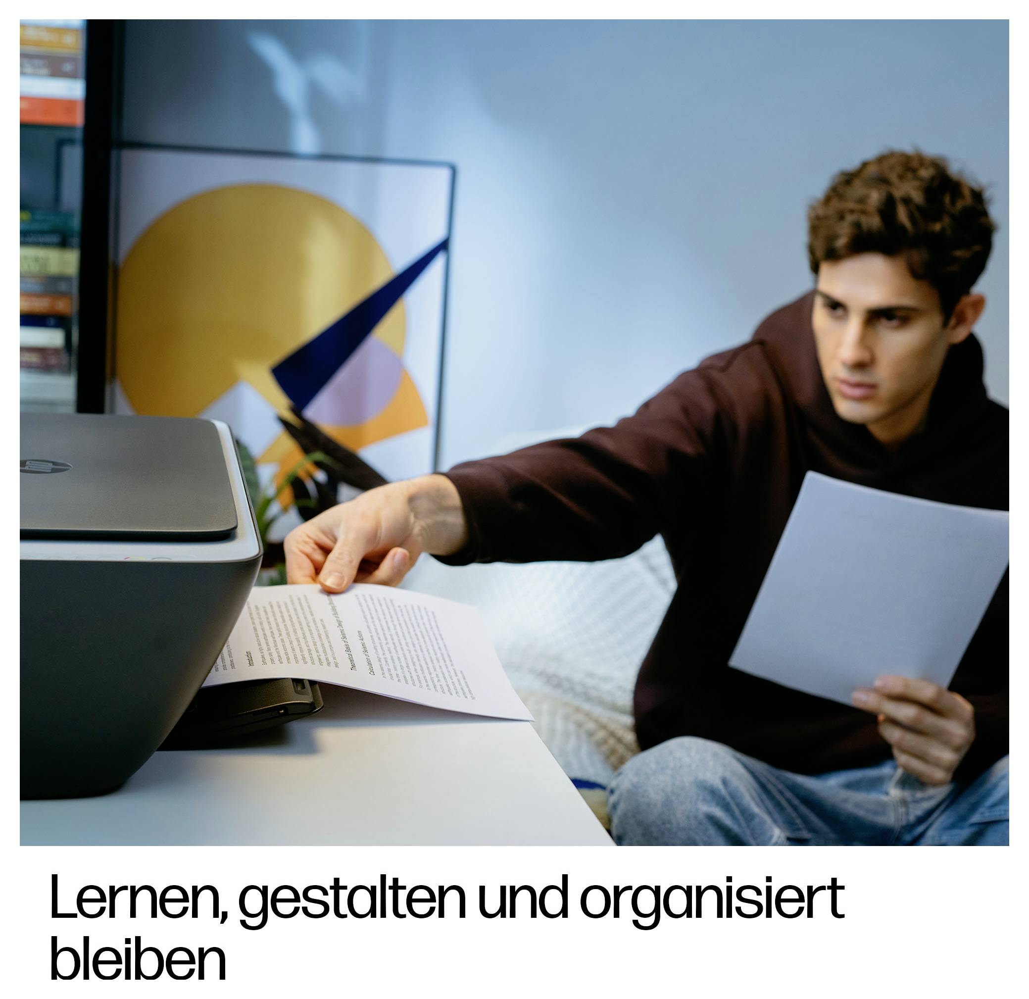 Mladý muž sedí u psacího stolu, drží kus papíru a podává jej do tiskárny. V pozadí je abstraktní umělecké dílo.