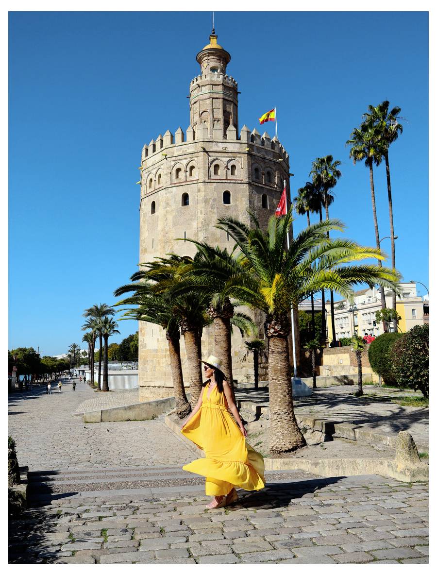 Žena v splývavých žlutých šatech kráčí poblíž Torre del Oro s palmami a jasně modrou oblohou v pozadí.