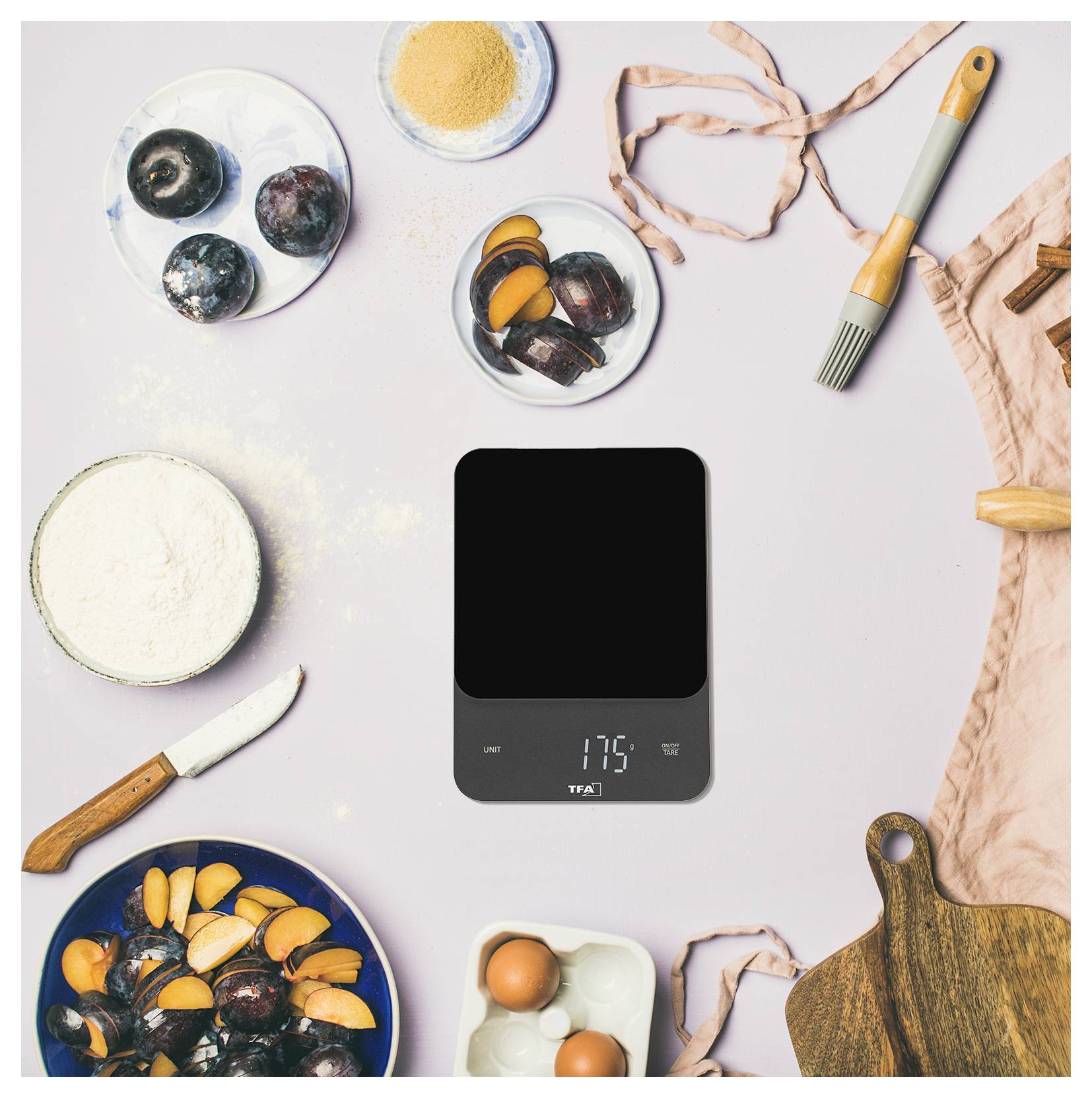 Pečicí ingredience a kuchyňské náčiní obklopují digitální kuchyňskou váhu zobrazující '175', včetně švestek, vajec, mouky, cukru a prkénka na krájení.