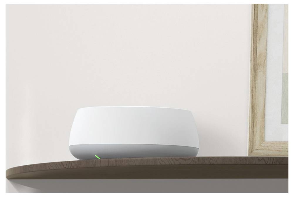 Moderní bílý router se zeleně svítícím světlem stojí na hnědé polici u jednoduché zdi vedle dřevěného rámovaného obrázku.