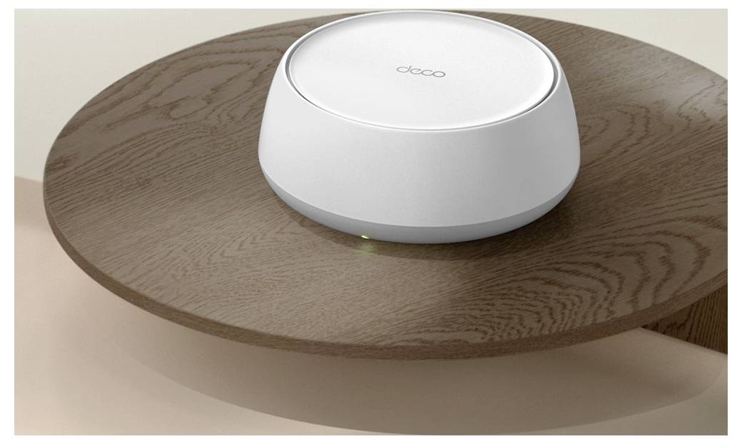 Bílý, kulatý bezdrátový router s jednoduchým designem leží na dřevěném stole a vpředu má malou zelenou kontrolní diodu.