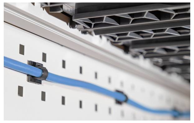 Modrý kabel je pečlivě připevněn k bílému perforovanému panelu, což naznačuje organizované vedení kabeláže v technickém prostředí.