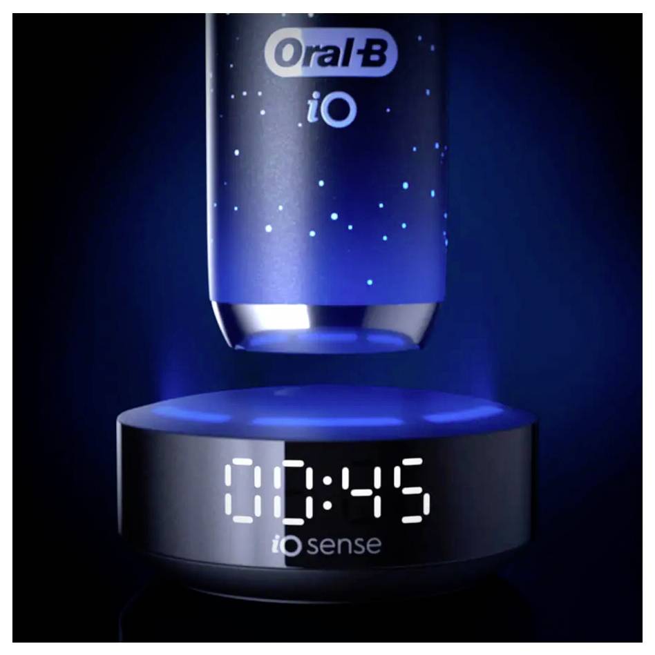 Elektrická zubní kartáček Oral-B iO vznášející se nad nabíjecí stanicí, s digitálními hodinami na základně zobrazujícími čas '10:45', obklopený modrým osvětlením.