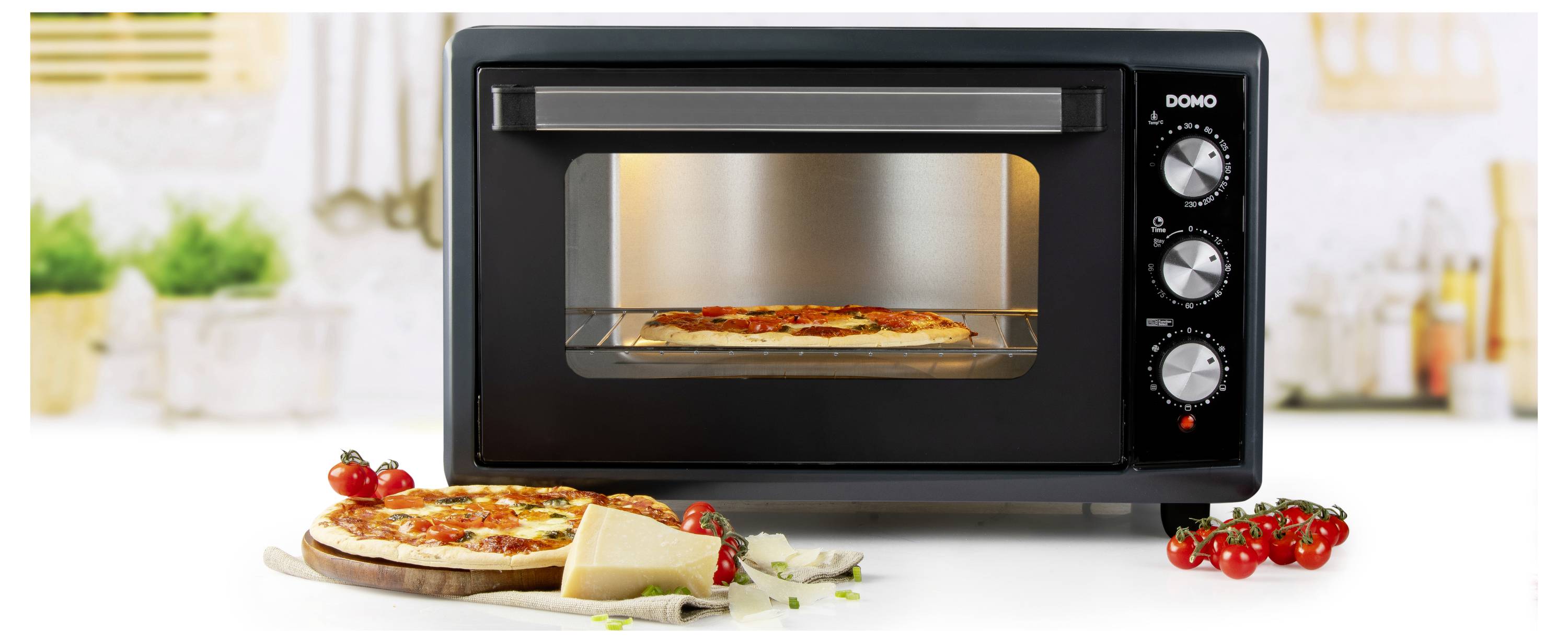 Pec na pizzu s pizzou uvnitř a další pizzou vedle ní, obklopená cherry rajčaty a sýrem, v prostředí kuchyně.